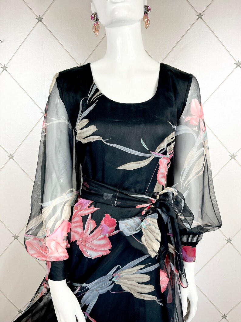 Peut inclure: Robe noire &agrave; imprim&eacute; floral avec manches transparentes et ceinture assortie. La robe pr&eacute;sente une encolure arrondie et une jupe fluide. Le motif floral comprend des accents roses, blancs et gris. La robe est pr&eacute;sent&eacute;e sur un mannequin.