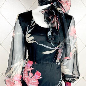 Peut inclure: Robe noire &agrave; imprim&eacute; floral avec manches transparentes et &eacute;charpe assortie nou&eacute;e en n&oelig;ud. La robe pr&eacute;sente une encolure arrondie et un corsage ajust&eacute;, avec une jupe fluide. Le motif floral comprend des accents roses, blancs et gris.