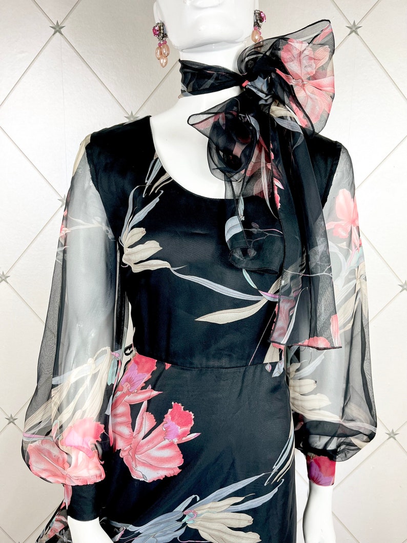 Peut inclure: Robe noire &agrave; imprim&eacute; floral avec manches transparentes et &eacute;charpe assortie nou&eacute;e en n&oelig;ud. La robe pr&eacute;sente une encolure arrondie et un corsage ajust&eacute;, avec une jupe fluide. Le motif floral comprend des accents roses, blancs et gris.