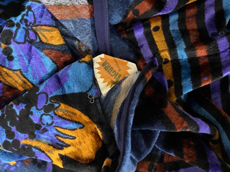 May include: A blue and purple velvet fabric with a floral pattern and a yellow tag that says "Floral" with the word "Exclusive" underneath. The fabric has a zipper and a black metal pull.