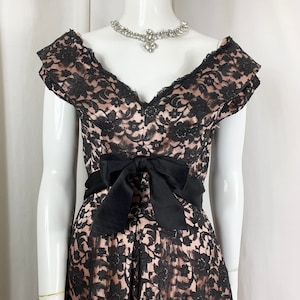May include: A black lace dress with a pink underlay. The dress has a V-neckline, short sleeves, and a black satin bow at the waist.