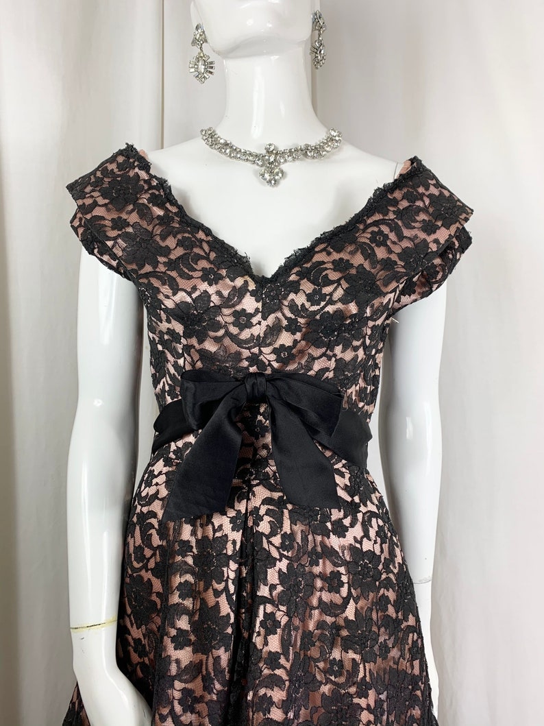 May include: A black lace dress with a pink underlay. The dress has a V-neckline, short sleeves, and a black satin bow at the waist.