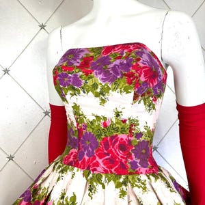 K&ouml;nnte beinhalten: Ein wei&szlig;es, tr&auml;gerloses Kleid mit vollem Rock und einem Blumendruck mit rosa, lila und gr&uuml;nen Blumen. Das Kleid hat ein tailliertes Oberteil und einen gerafften Rock.