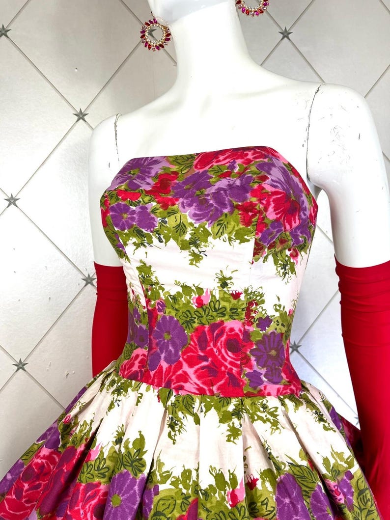 K&ouml;nnte beinhalten: Ein wei&szlig;es, tr&auml;gerloses Kleid mit vollem Rock und einem Blumendruck mit rosa, lila und gr&uuml;nen Blumen. Das Kleid hat ein tailliertes Oberteil und einen gerafften Rock.