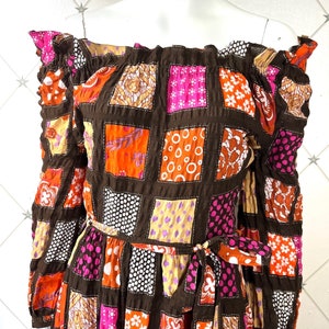 K&ouml;nnte beinhalten: Schulterfreies Kleid mit langen &Auml;rmeln und Patchwork-Muster. Das Kleid hat einen braunen Hintergrund mit Quadraten aus orangefarbenem, rosa, wei&szlig;em und violettem Stoff. Das Kleid hat einen G&uuml;rtel an der Taille.