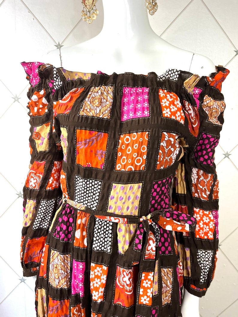 K&ouml;nnte beinhalten: Schulterfreies Kleid mit langen &Auml;rmeln und Patchwork-Muster. Das Kleid hat einen braunen Hintergrund mit Quadraten aus orangefarbenem, rosa, wei&szlig;em und violettem Stoff. Das Kleid hat einen G&uuml;rtel an der Taille.