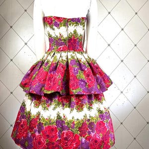 K&ouml;nnte beinhalten: Ein wei&szlig;es tr&auml;gerloses Kleid mit einem mehrlagigen Rock. Das Kleid ist mit einem Blumendruck mit rosa, lila und gr&uuml;nen Blumen bedeckt.