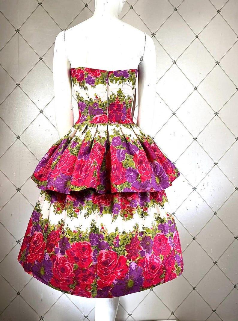 K&ouml;nnte beinhalten: Ein wei&szlig;es tr&auml;gerloses Kleid mit einem mehrlagigen Rock. Das Kleid ist mit einem Blumendruck mit rosa, lila und gr&uuml;nen Blumen bedeckt.