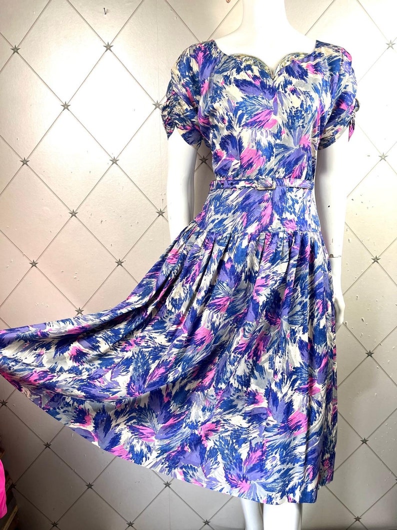 May include: A vintage floral dress with a white background and blue, pink, and purple abstract floral print. The dress has a sweetheart neckline, short sleeves, and a fitted waist with a belt.