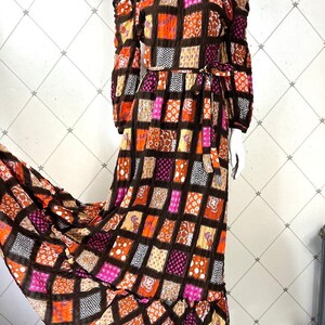 K&ouml;nnte beinhalten: Ein langes, schulterfreies Maxikleid mit Patchwork-Muster. Das Kleid hat einen braunen Hintergrund mit orangen, rosa und wei&szlig;en Quadraten. Das Kleid hat einen G&uuml;rtel in der Taille und einen R&uuml;schen-Saum.