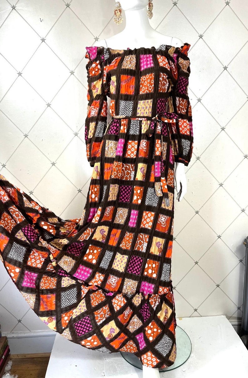 K&ouml;nnte beinhalten: Ein langes, schulterfreies Maxikleid mit Patchwork-Muster. Das Kleid hat einen braunen Hintergrund mit orangen, rosa und wei&szlig;en Quadraten. Das Kleid hat einen G&uuml;rtel in der Taille und einen R&uuml;schen-Saum.