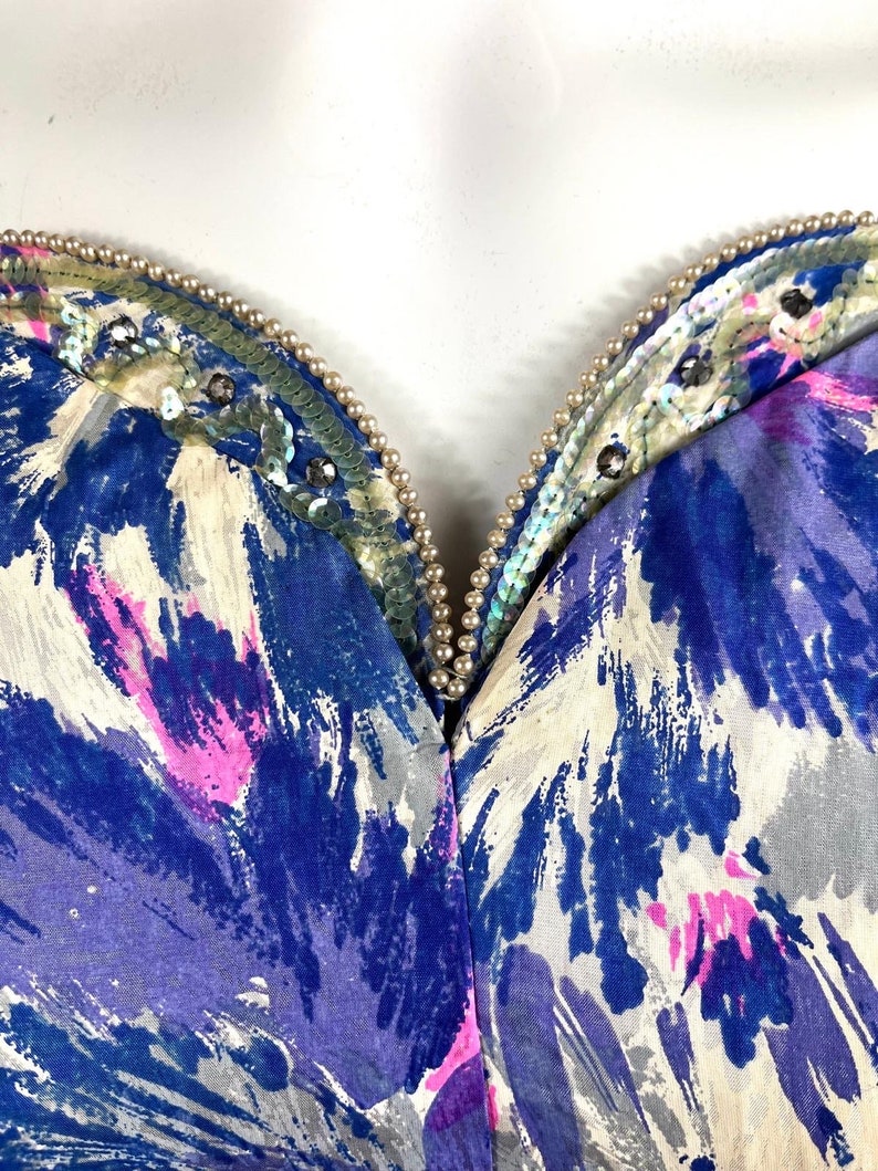 May include: A blue, pink, and white abstract print fabric with a beaded neckline. The neckline is adorned with iridescent sequins and small white pearls.