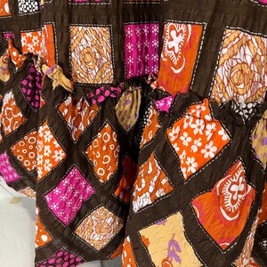 K&ouml;nnte beinhalten: Ein brauner Rock mit Patchwork-Muster aus Quadraten aus orangefarbenem, rosa und wei&szlig;em Stoff mit verschiedenen Mustern. Der Rock hat einen R&uuml;schen-Saum.