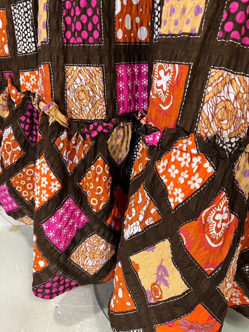 K&ouml;nnte beinhalten: Ein brauner Rock mit Patchwork-Muster aus Quadraten aus orangefarbenem, rosa und wei&szlig;em Stoff mit verschiedenen Mustern. Der Rock hat einen R&uuml;schen-Saum.
