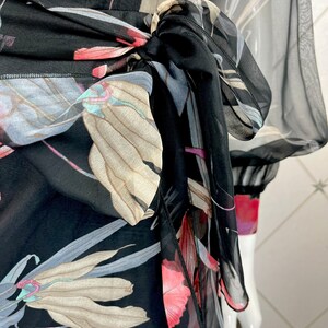 Peut inclure: Robe noire en tissu transparent avec un motif floral rose, beige et gris. La robe est nou&eacute;e &agrave; la taille et a des manches longues. Le motif floral est visible sur toute la robe.