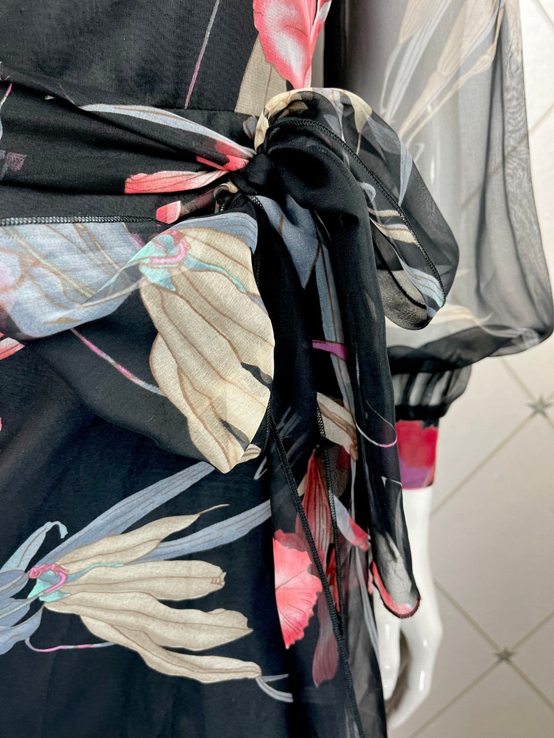Peut inclure: Robe noire en tissu transparent avec un motif floral rose, beige et gris. La robe est nou&eacute;e &agrave; la taille et a des manches longues. Le motif floral est visible sur toute la robe.