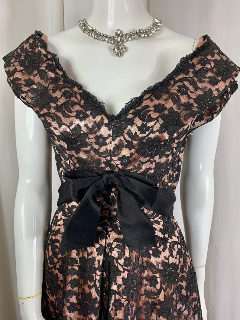 May include: A black and blush pink lace dress with a black satin bow at the waist. The dress has a V-neckline and short sleeves.