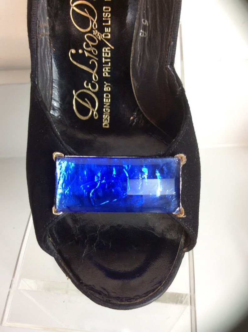 Puede incluir: Zapato de tac&oacute;n alto negro con punta abierta y un gran adorno rectangular de joya azul. El zapato est&aacute; hecho de ante y cuero, con letras doradas en el interior que dicen "De Liso Deb".