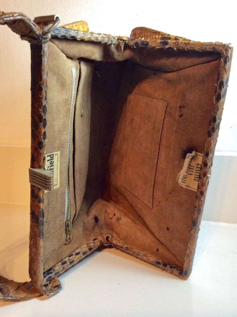 May include: Inside view of a vintage snakeskin handbag with a zippered pocket and a label that reads "Field". The bag has a brown suede lining and a small pocket on the opposite side of the zippered pocket. The label on the small pocket reads "Tailored  Exclusively for  Jay's".