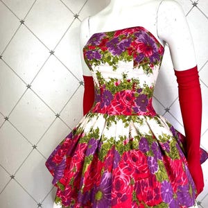 K&ouml;nnte beinhalten: Ein wei&szlig;es tr&auml;gerloses Kleid mit einem Stufenrock und einem Blumendruck mit rosa, lila und gr&uuml;nen Rosen. Das Kleid befindet sich auf einer wei&szlig;en Puppe mit roten Handschuhen.
