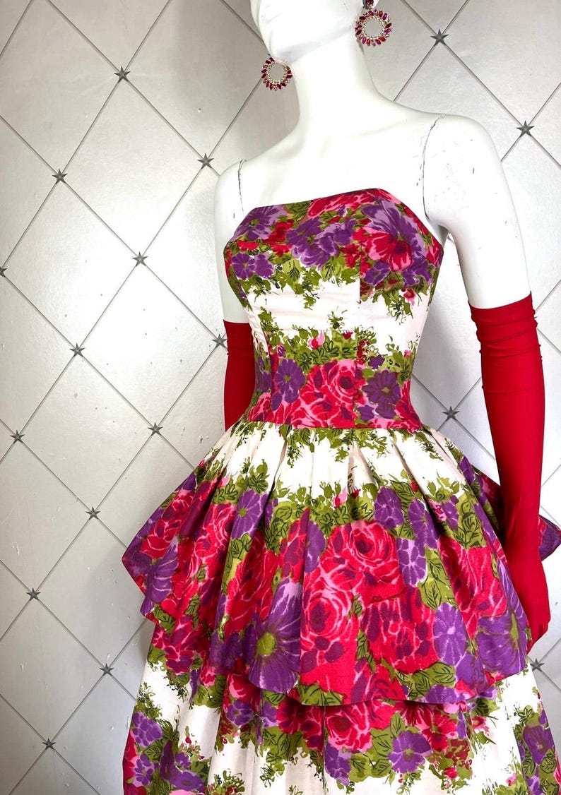 K&ouml;nnte beinhalten: Ein wei&szlig;es tr&auml;gerloses Kleid mit einem Stufenrock und einem Blumendruck mit rosa, lila und gr&uuml;nen Rosen. Das Kleid befindet sich auf einer wei&szlig;en Puppe mit roten Handschuhen.