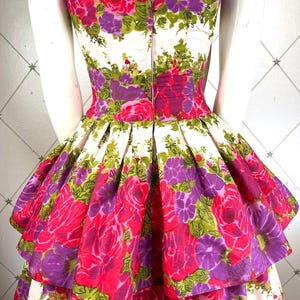 K&ouml;nnte beinhalten: Ein tr&auml;gerloses Kleid mit wei&szlig;em Hintergrund und einem pink- und lilafarbenen Blumendruck. Das Kleid hat einen mehrlagigen Rock mit einem gerafften Saum.