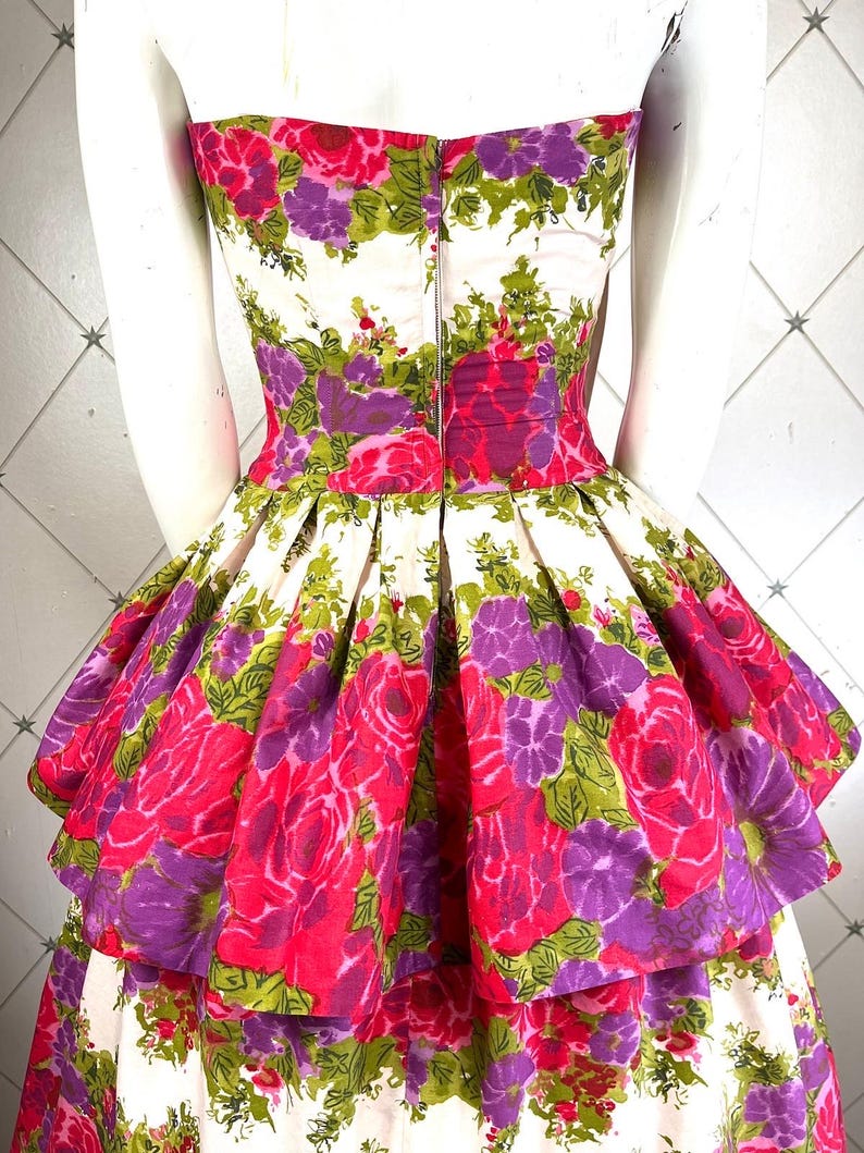 K&ouml;nnte beinhalten: Ein tr&auml;gerloses Kleid mit wei&szlig;em Hintergrund und einem pink- und lilafarbenen Blumendruck. Das Kleid hat einen mehrlagigen Rock mit einem gerafften Saum.