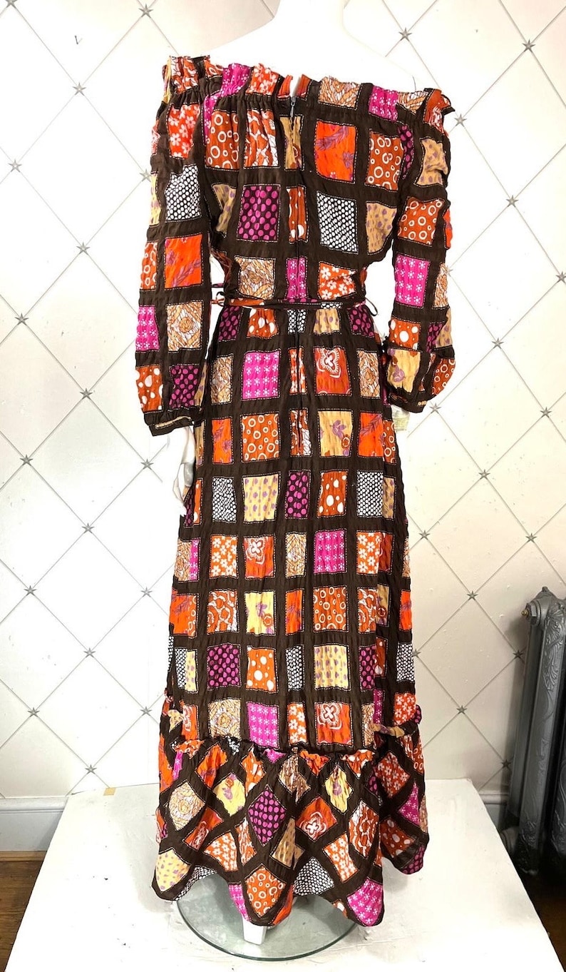 K&ouml;nnte beinhalten: Ein langes, schulterfreies Kleid mit Patchwork-Muster. Das Kleid ist aus braunem, orangefarbenem, rosa und gelbem Stoff. Der Stoff hat eine gesteppte Textur. Das Kleid hat lange &Auml;rmel und ein tailliertes Oberteil. Der Rock ist weit und flie&szlig;end.