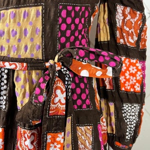 K&ouml;nnte beinhalten: Ein braunes Kleid mit Patchwork-Muster, das orange, rosa, wei&szlig;e und braune Blumenmuster zeigt. Das Kleid hat lange &Auml;rmel und eine Schleife in der Taille.