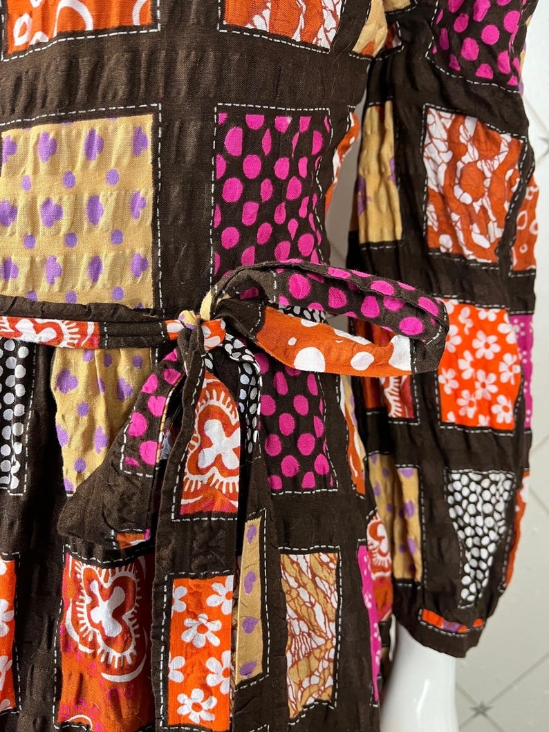 K&ouml;nnte beinhalten: Ein braunes Kleid mit Patchwork-Muster, das orange, rosa, wei&szlig;e und braune Blumenmuster zeigt. Das Kleid hat lange &Auml;rmel und eine Schleife in der Taille.