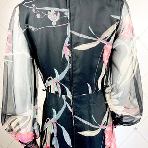 Peut inclure: Robe noire &agrave; imprim&eacute; floral avec manches longues transparentes. La robe pr&eacute;sente un motif floral aux tons rose, blanc et vert. Elle a un col montant et un corsage ajust&eacute;, avec une jupe fluide.