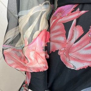 Peut inclure: Robe noire avec un motif floral dans les tons de rose, rouge et beige. La robe a de longues manches transparentes avec un poignet noir. Le motif floral est visible sur les manches et le corps de la robe.