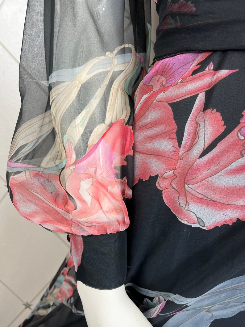 Peut inclure: Robe noire avec un motif floral dans les tons de rose, rouge et beige. La robe a de longues manches transparentes avec un poignet noir. Le motif floral est visible sur les manches et le corps de la robe.