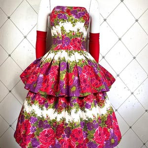 K&ouml;nnte beinhalten: Ein tr&auml;gerloses Kleid mit wei&szlig;em Hintergrund und einem Blumendruck in Rosa-, Lila- und Gr&uuml;nt&ouml;nen. Das Kleid hat einen Stufenrock und ist am Oberteil figurbetont.