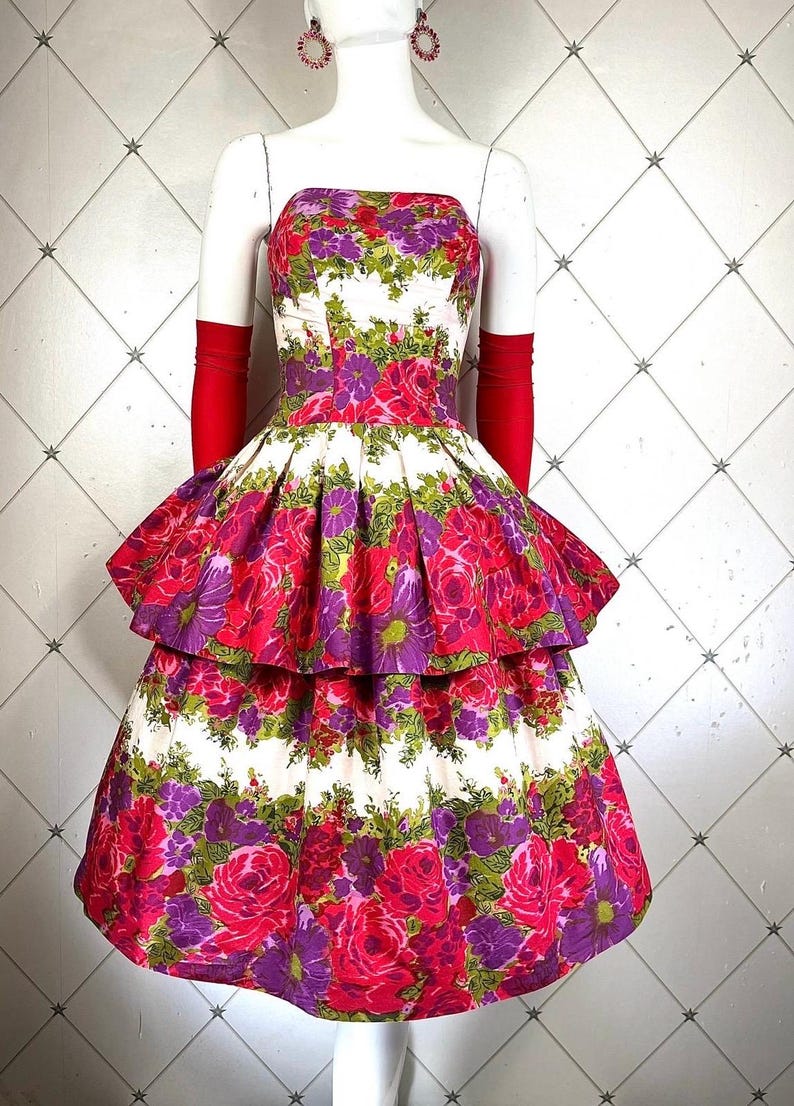 K&ouml;nnte beinhalten: Ein tr&auml;gerloses Kleid mit wei&szlig;em Hintergrund und einem Blumendruck in Rosa-, Lila- und Gr&uuml;nt&ouml;nen. Das Kleid hat einen Stufenrock und ist am Oberteil figurbetont.