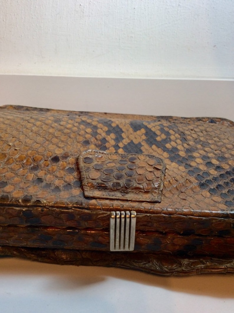 May include: A vintage brown snakeskin clutch purse with a silver clasp. The purse has a textured surface and a flap closure.
