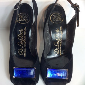 Puede incluir: Un par de zapatos de tac&oacute;n negros con correa trasera y una joya rectangular azul en la puntera. Los tacones tienen un cierre de hebilla y la marca "Dolcis" es visible en la plantilla. Tambi&eacute;n es visible el texto "De Liso Debs".