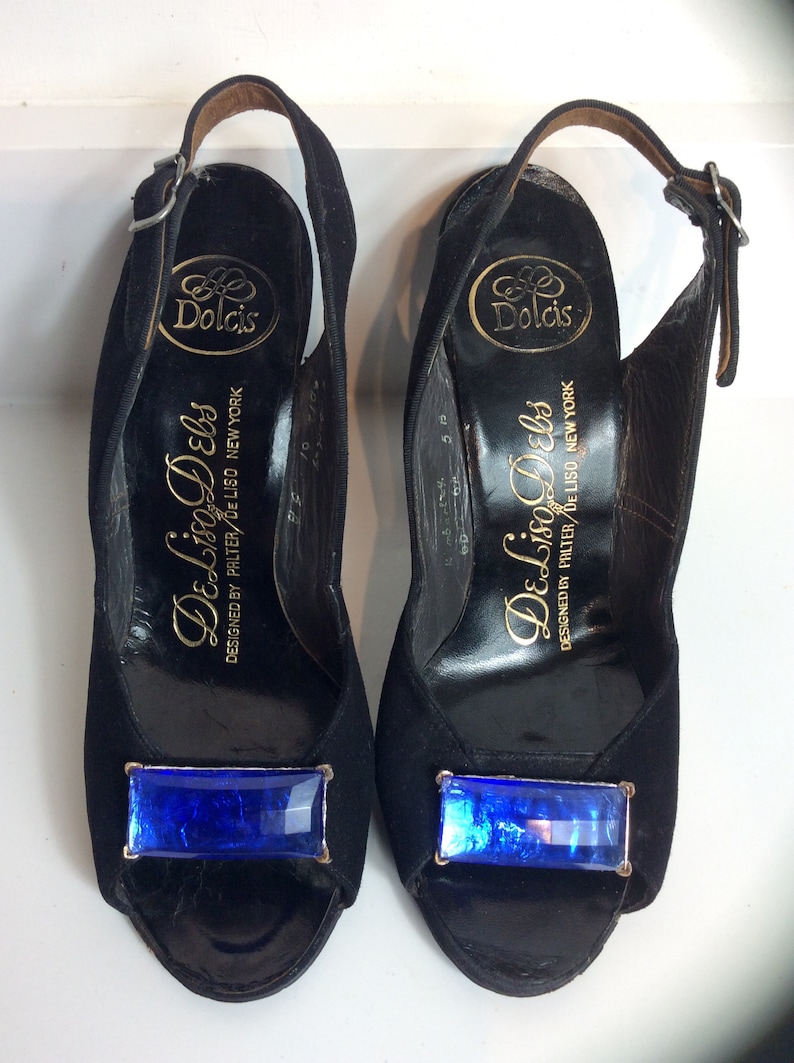 Puede incluir: Un par de zapatos de tac&oacute;n negros con correa trasera y una joya rectangular azul en la puntera. Los tacones tienen un cierre de hebilla y la marca "Dolcis" es visible en la plantilla. Tambi&eacute;n es visible el texto "De Liso Debs".