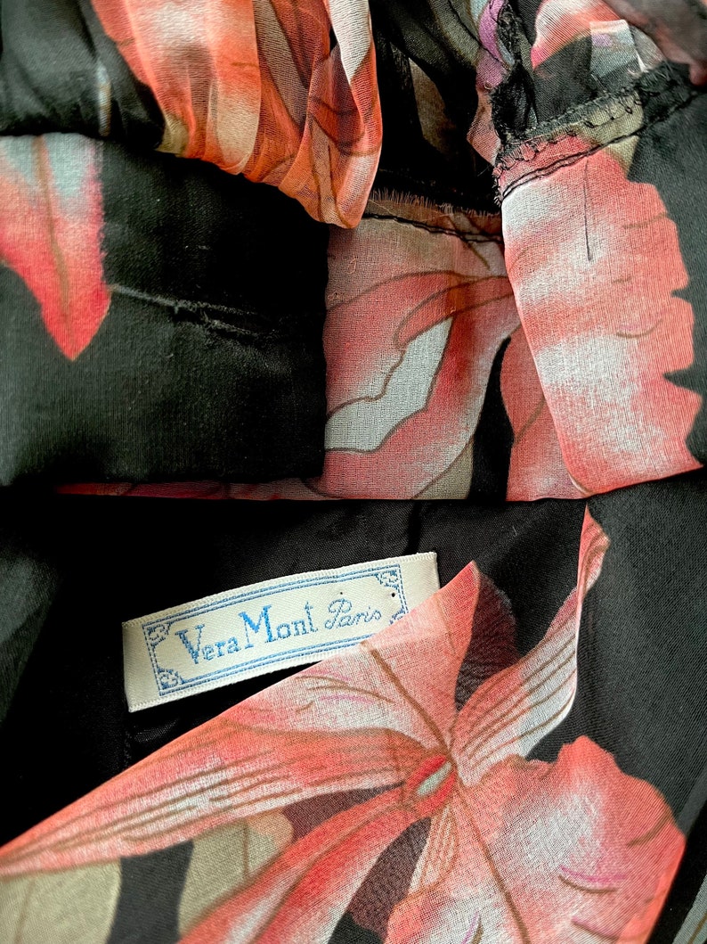 Peut inclure: Gros plan sur un v&ecirc;tement noir avec un motif floral dans les tons de rose, de rouge et de blanc. Le tissu est transparent et superpos&eacute;. Une &eacute;tiquette avec le texte "Vera Mont Paris" est visible.