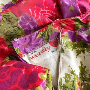 K&ouml;nnte beinhalten: Ein wei&szlig;es Kleid mit einem rosa-violetten Blumendruck. Das Kleid hat einen Rei&szlig;verschluss und ein Etikett mit der Aufschrift "Your Wendy Dress".