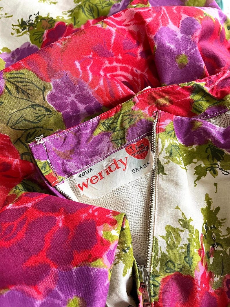K&ouml;nnte beinhalten: Ein wei&szlig;es Kleid mit einem rosa-violetten Blumendruck. Das Kleid hat einen Rei&szlig;verschluss und ein Etikett mit der Aufschrift "Your Wendy Dress".
