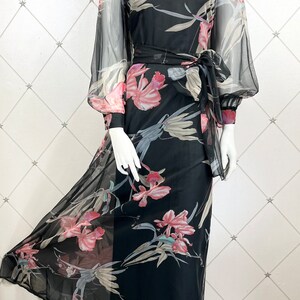 Peut inclure: Longue robe noire &agrave; imprim&eacute; floral, avec de longues manches transparentes et une ceinture &agrave; nouer assortie. La robe pr&eacute;sente une encolure arrondie et une jupe fluide. Le motif floral comprend des fleurs roses et blanches et des feuilles vertes.