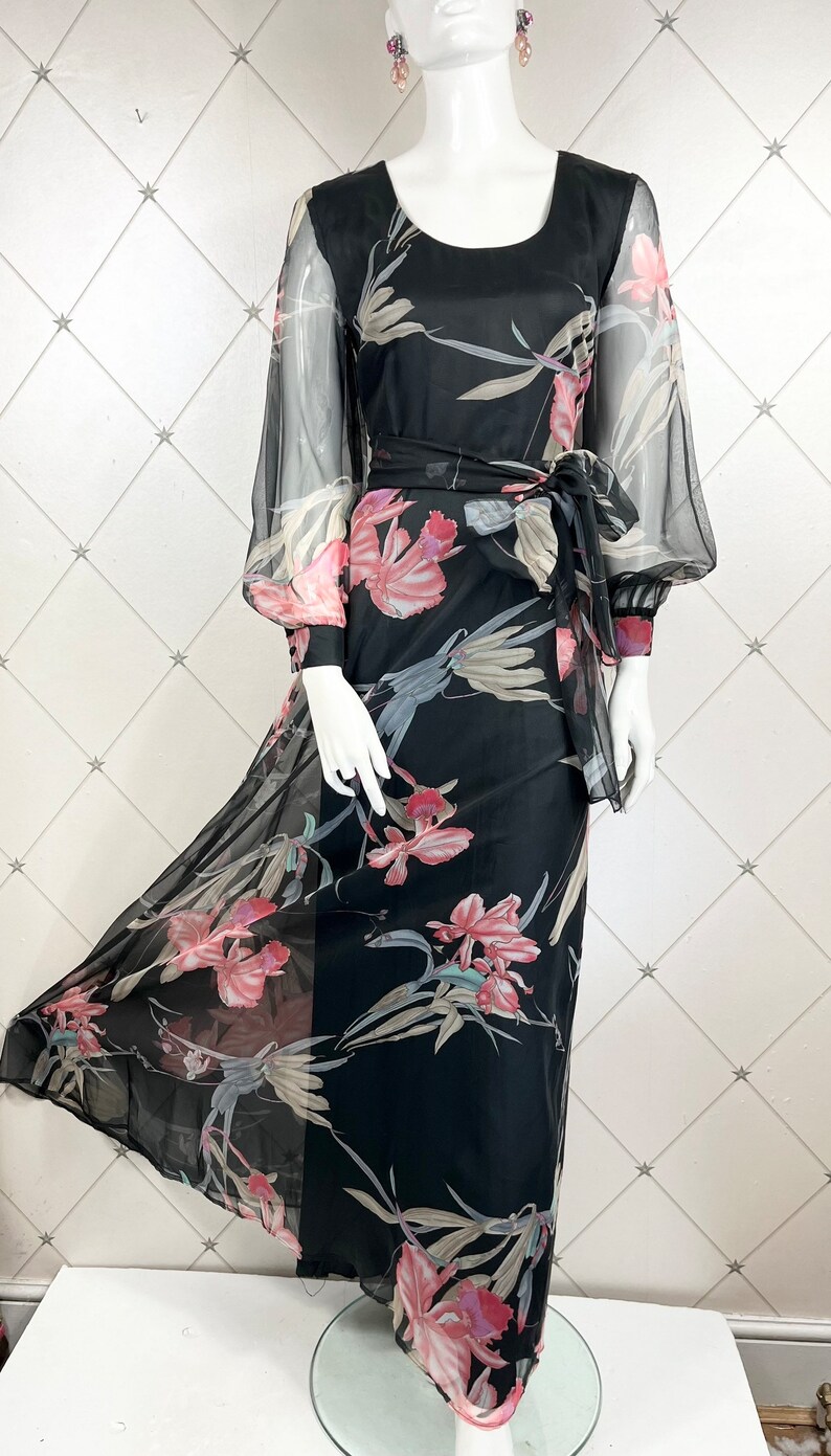 Peut inclure: Longue robe noire &agrave; imprim&eacute; floral, avec de longues manches transparentes et une ceinture &agrave; nouer assortie. La robe pr&eacute;sente une encolure arrondie et une jupe fluide. Le motif floral comprend des fleurs roses et blanches et des feuilles vertes.