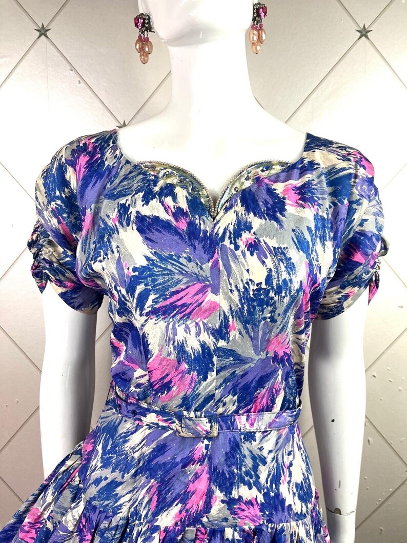 May include: A vintage dress with a blue, pink, and white abstract floral print. The dress has a sweetheart neckline, short sleeves, and a fitted bodice. The dress is cinched at the waist with a matching belt.