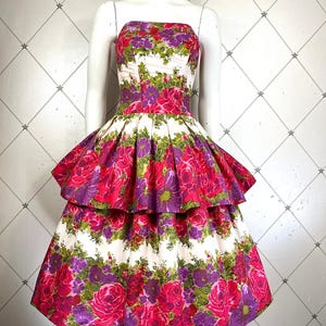 K&ouml;nnte beinhalten: Ein wei&szlig;es, tr&auml;gerloses Kleid mit einem pink- und lilafarbenen Blumendruck. Das Kleid hat einen Stufenrock mit einem R&uuml;schen-Saum.