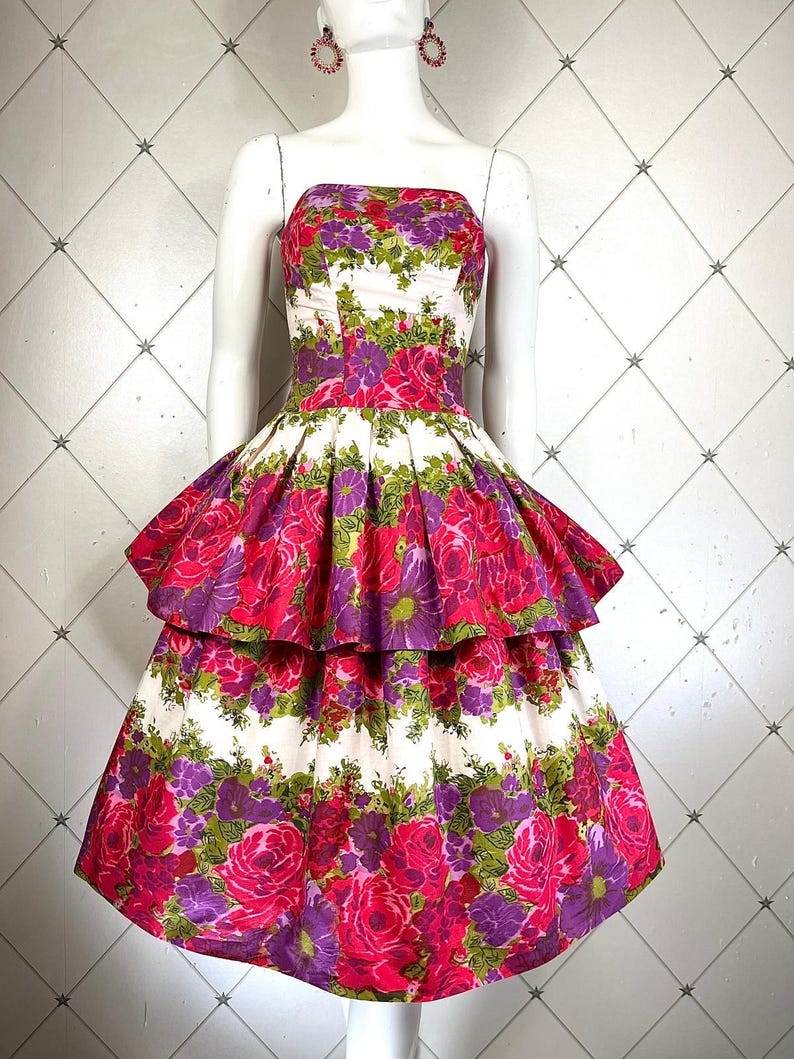 K&ouml;nnte beinhalten: Ein wei&szlig;es, tr&auml;gerloses Kleid mit einem pink- und lilafarbenen Blumendruck. Das Kleid hat einen Stufenrock mit einem R&uuml;schen-Saum.