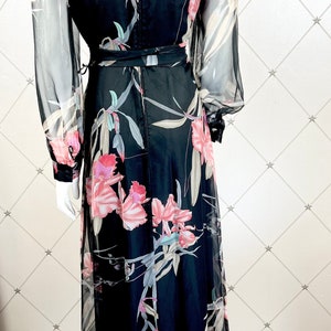 Peut inclure: Longue robe noire avec un imprim&eacute; floral d'orchid&eacute;es roses et blanches. La robe a de longues manches transparentes et une ceinture assortie. Le dos de la robe a une fermeture &eacute;clair.