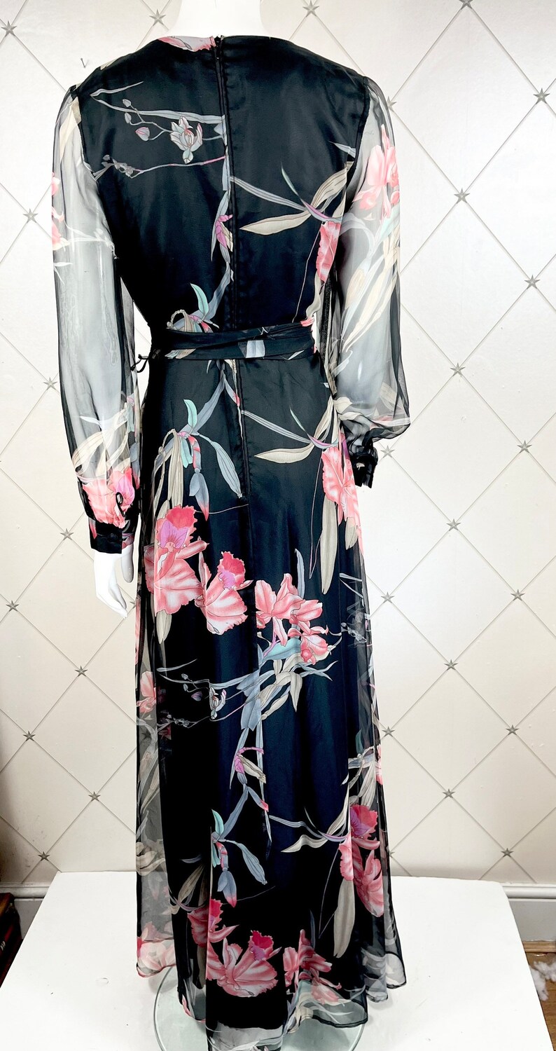 Peut inclure: Longue robe noire avec un imprim&eacute; floral d'orchid&eacute;es roses et blanches. La robe a de longues manches transparentes et une ceinture assortie. Le dos de la robe a une fermeture &eacute;clair.