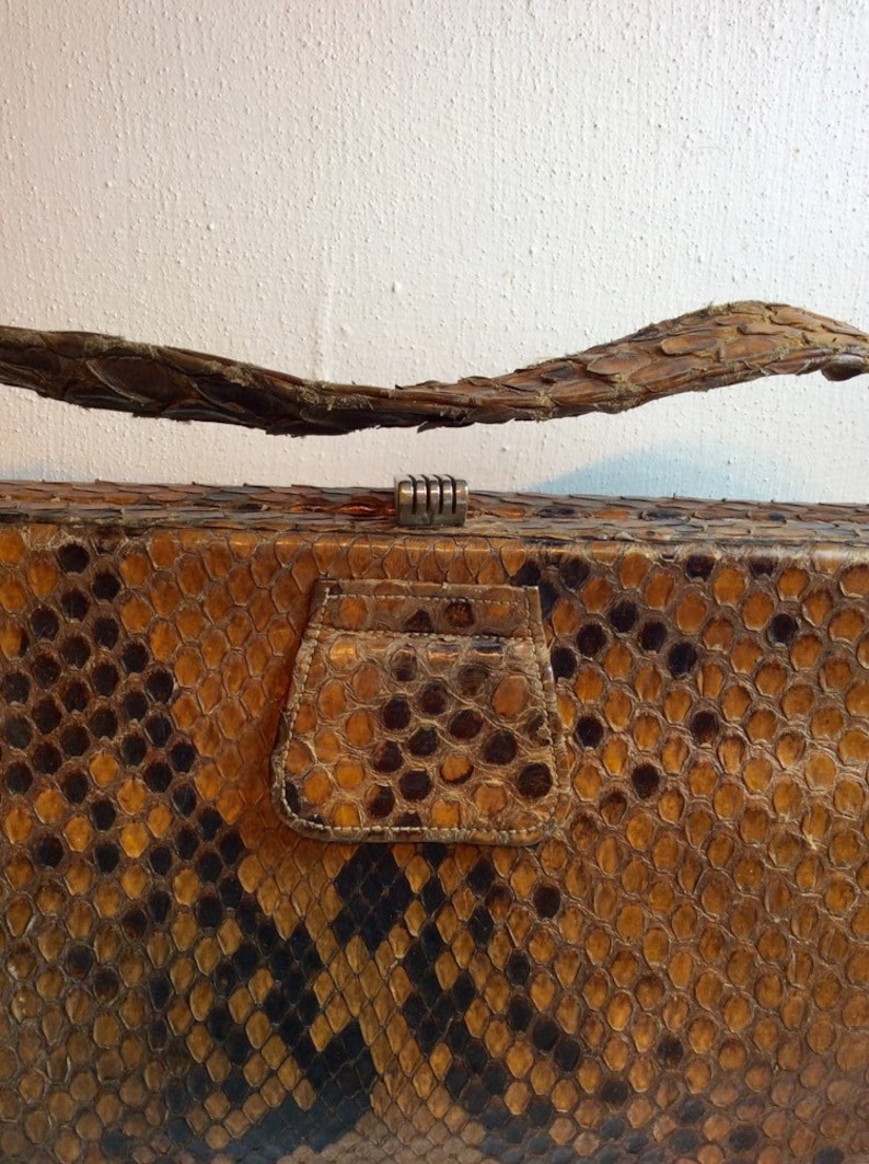 May include: A vintage snakeskin clutch purse with a brown and black pattern. The purse has a metal clasp and a flap with a small pocket.