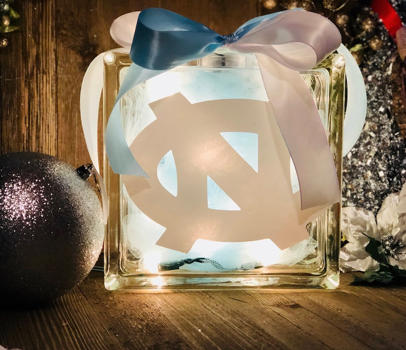 May include: A glass block with a white UNC logo and a blue and white ribbon bow. The block is illuminated from within, creating a warm glow.